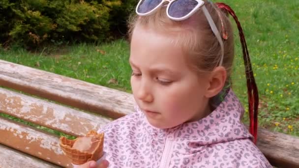 可愛い女の子がベンチの屋外でワッフルコーンでアイスクリームを食べています サングラスの子供は晴れた日に緑の公園でチョコレートジェラートイタリアの冷凍デザートをかむ 甘い冷たいおいしいさわやかなシャーベット — ストック動画