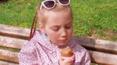 Pretty little girl eating an ice cream in waffle cone on bench outdoor. Child in sunglasses bites chocolate gelato italian frozen dessert in green park on sunny day. Sweet cold tasty refreshing sorbet