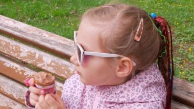 Pretty little girl eating an ice cream in waffle cone on bench outdoor. Child in sunglasses bites chocolate gelato italian frozen dessert in green park on sunny day. Sweet cold tasty refreshing sorbet