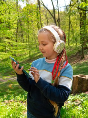 Adorable child girl in headphones learning language listening to music audio books podcasts smartphone outdoor. Blogger kid with gadget distance school online lesson chat cell phone conference in park