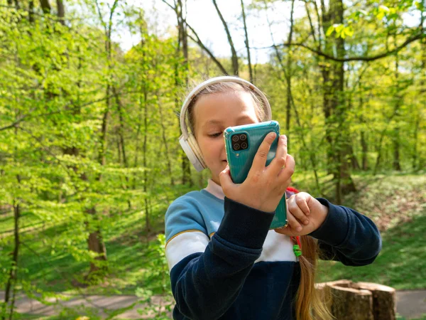 可爱的小女孩在耳机学习语言听音乐有声书籍播客智能手机室外 在公园里 带着小玩艺儿远程学校在线课上聊天手机会议的博客孩子 — 图库照片