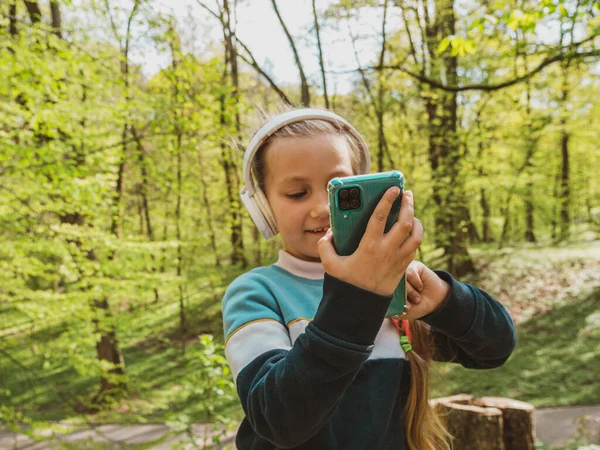 可爱的小女孩在耳机学习语言听音乐使用智能手机室外晴天 在公园里 带着小玩艺儿的博客孩子在线上课时聊天 — 图库照片