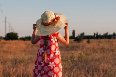 Hasır şapkalı çocuk buğday tarlasında elbise giyiyor. Gün batımında doğa manzarasına bakan sevimli bir çocuk. Aile tarımı tarım çevre ekolojisi kavramı. Cottagecore tarzı estetik.