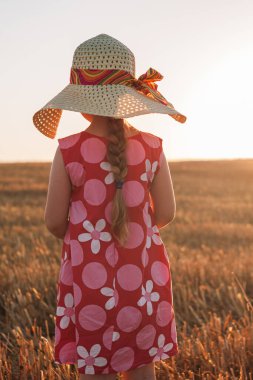 Hasır şapkalı çocuk buğday tarlasında elbise giyiyor. Gün batımında doğa manzarasına bakan sevimli bir çocuk. Aile tarımı tarım çevre ekolojisi kavramı. Cottagecore tarzı estetik.
