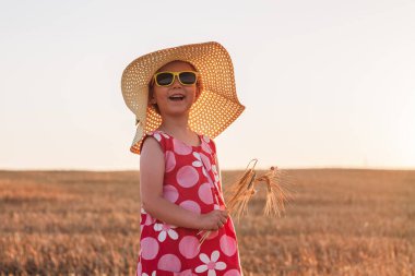 Hasır şapkalı çocuk buğday tarlasında elbise giyiyor. Gün batımında kırsal alanda güneş gözlüklü gülümseyen bir çocuk. Aile tarımı tarım çevre ekolojisi kavramı. Cottagecore tarzı estetik.