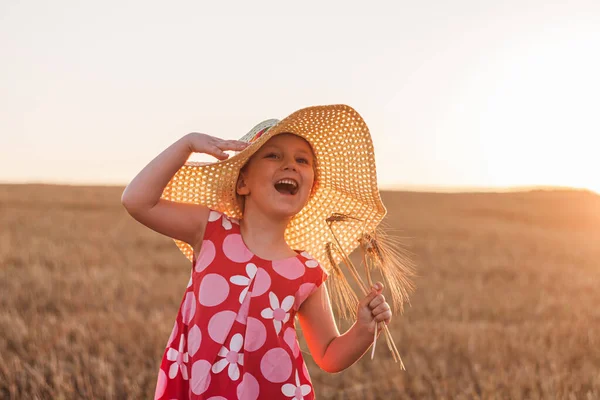 Child girl in straw hat dress in wheat spikelets field. Smiling kid in sunglasses on sunset countryside landscape. Family farming agriculture environment ecology concept. Cottagecore style aesthetic.