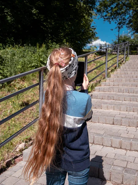 可爱的小女孩 长发在社交媒体的智能手机里聊天 在公园的楼梯上边打电话边看书 带小玩艺儿的博客小子在网上直播手机短信 — 图库照片
