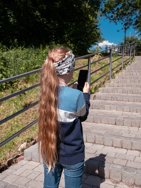 可爱的小女孩 长发在社交媒体的智能手机里聊天 在公园的楼梯上边打电话边看书 带小玩艺儿的博客小子在网上直播手机短信 — 图库照片