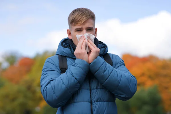 Tampan pria meniup hidungnya dalam saputangan kertas luar ruangan di alam, emas musim gugur latar belakang. Potret orang sakit muda dengan hidung beringus, flu. Gejala dari coronavirus, covid-19. Alergi — Stok Foto