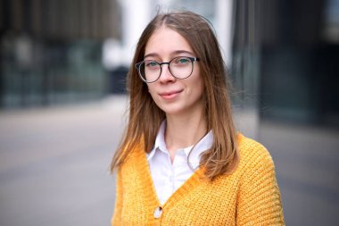 Güzel genç bir kızın portresi, gözlüklü bir öğrenci, kameraya bakıyor, açık hava üniversitesinde ya da üniversitede durup gülümsüyor.