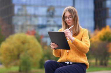 Güzel genç üniversite ya da gözlüklü üniversiteli kız parkta oturuyor, üniversite kampüsünde, elektronik kalem ya da kalemle tablet bilgisayarına resim çiziyor, cihazıyla internetten çalışıyor.
