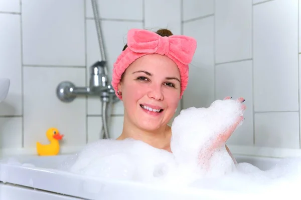 Beauty portrait of beautiful happy positive girl, young cheerful woman is relaxing, lying in bathroom, taking a bath with bubbles, smiling and looking at camera, washing her body with foam in bathtub