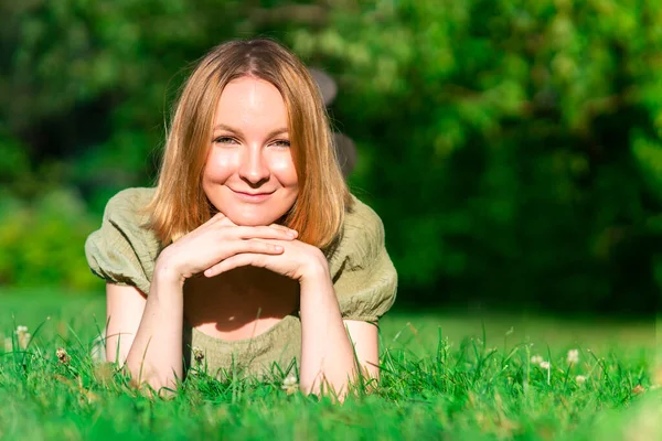 Портрет молодой счастливой веселой позитивной девочки-подростка, молодой, довольно радостной женщины лежит на желудке на зеленой траве, улыбается, смотрит в камеру, наслаждается летним добрым днем. Копирование пространства, место для текста — стоковое фото