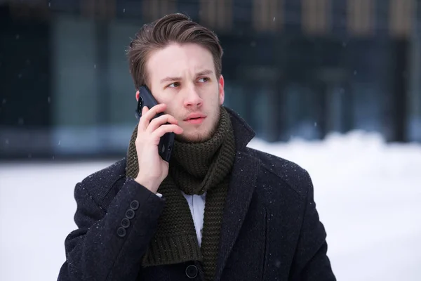Portrait of handsome upset unhappy European blonde guy young nervous man, businessman is talking at cell mobile phone calling on smartphone with serious face outdoors at cold winter snowy day — Stock Photo, Image
