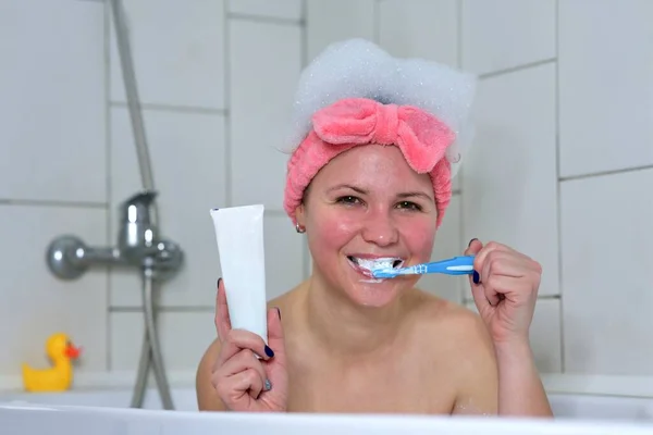 Портрет счастливой веселой позитивной красивой девушки, молодая женщина чистит свои здоровые белые зубы с улыбкой зубной щетки, принимая ванну в ванной комнате. Тюбик зубной пасты с копировальным пространством, макет — стоковое фото