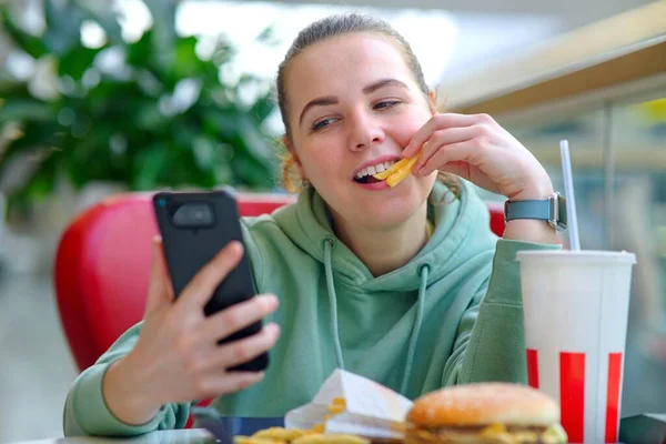 Joven mujer hermosa, feliz chica positiva comiendo comida chatarra rápida: hamburguesa, hamburguesa, papas fritas y refrescos en el patio de comidas en el centro comercial y utilizando la mirada en su teléfono móvil, teléfono inteligente, sonriendo — Foto de Stock
