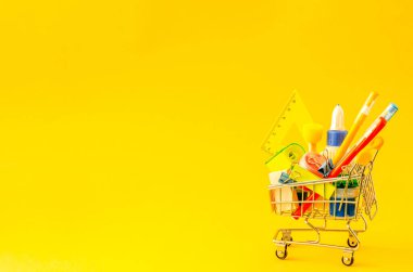 Assorted stationery in a shopping trolley on a yellow background with place for text. School preparation concept, flat lay, copy space. View from above.