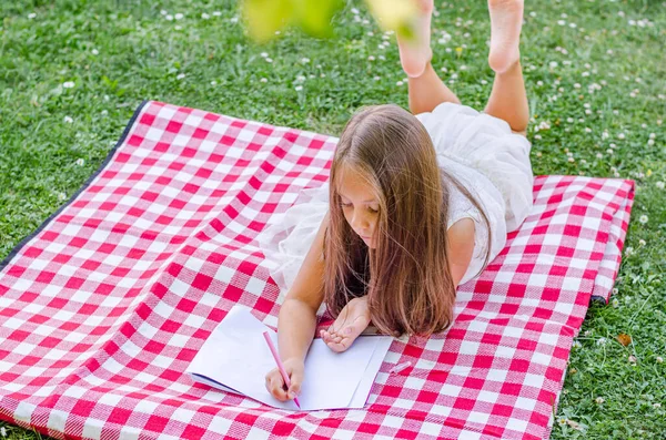 The girl lies on the grass in the garden under a tree in summer and writes on paper with a pen. The concept of seasonality, recreation, child development. Close-up.
