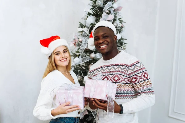 Jóvenes parejas multinacionales en los sombreros de Santa tienen regalos en el fondo de un árbol de Navidad y sonríen mientras miran a la cámara. Concepto de Navidad y Año Nuevo —  Fotos de Stock
