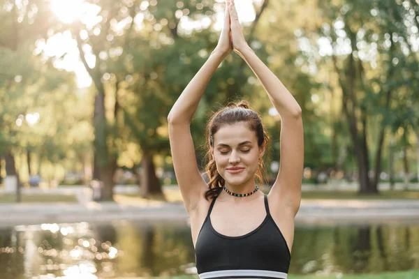 Ung kvinna utövar yoga i morgonparken nära sjön. Träning utomhus. Självsäker ung kaukasiska kvinna i sport topp tittar åt sidan och håller ut sina armar i en grön park på en solig — Stockfoto
