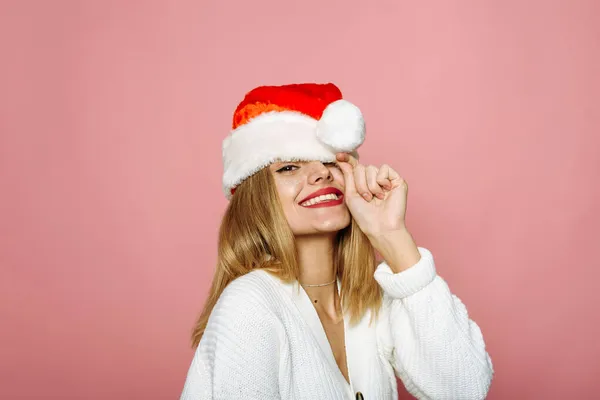 Una mujer divertida cubre un ojo con un sombrero de Santa Claus y se ríe. La dama coqueta sobre el fondo rosado celebra la Navidad y el Año Nuevo —  Fotos de Stock