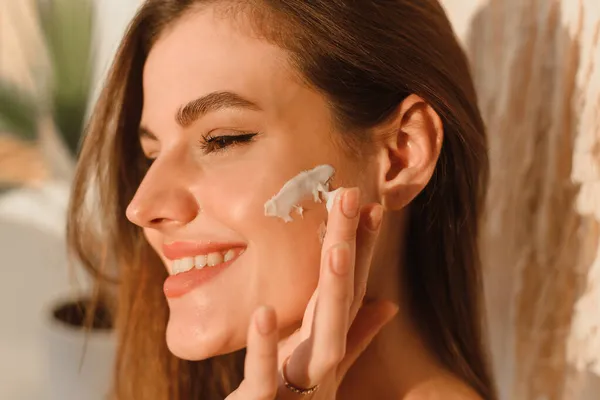 Una mujer joven se aplica crema hidratante en la cara en su baño de sol. Concepto de rutina diaria, hidratación y cuidado de la piel — Foto de Stock