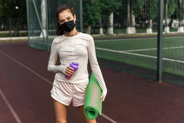 Een jonge vrouw met een zwart beschermend masker staat op het sportterrein en houdt een yogamat en een fles water vast. Sportactiviteiten tijdens quarantaine. Buiten — Stockfoto