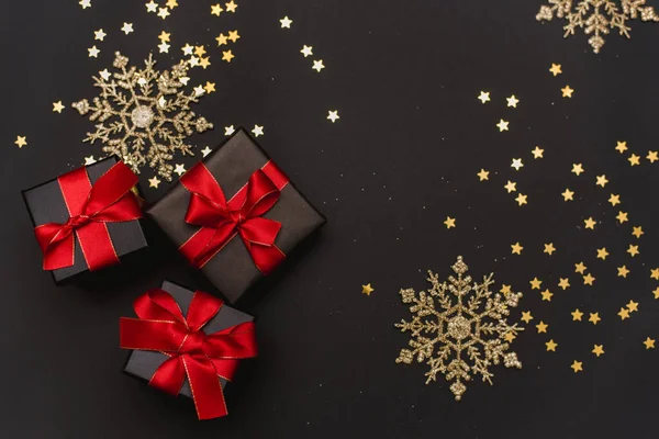 Zwarte geschenkdozen met een rood lint op een gouden confetti achtergrond. Elegante kerstachtergrond met moderne gouden kerstversieringen, ballen, geschenkdozen, confetti sterren op donkerzwarte achtergrond — Stockfoto