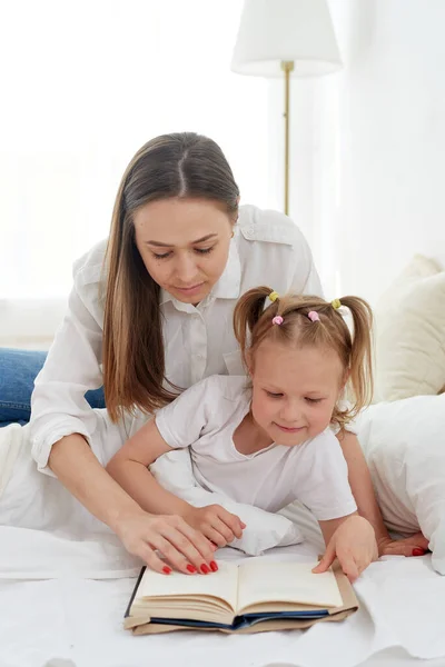 Close-up mom and little daughter are resting in a cozy bed, reading a funny interesting childrens book together — Stock Photo, Image