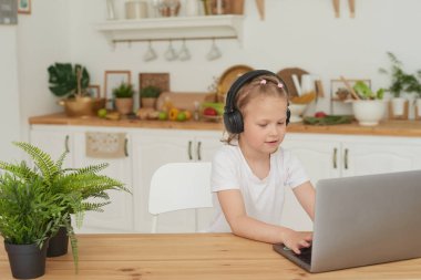 Çocuklar için çevrimiçi eğitim. Liseli kız evdeki bilgisayarlı görüntülü sohbetten video konferans üzerinden online ders izliyor. Küçük kız mutfakta oturmuş kulaklıkla oyun oynuyor.