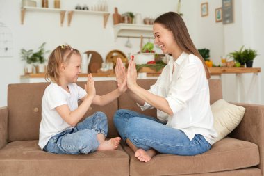Positive cheerful mom and her daughter spend the weekend together, sit on the couch, play pat a cake in the home room indoors