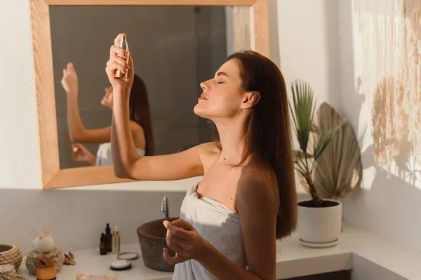 Una hermosa joven, envuelta en una toalla, se rocía agua mineral en la cara frente a un espejo. Concepto de hidratación cutánea. — Foto de Stock