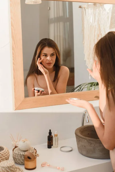 Una mujer joven se aplica crema hidratante en la cara en su baño de sol. Concepto de rutina diaria, hidratación y cuidado de la piel — Foto de Stock