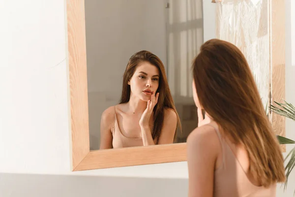 Attractive smiling young brunette looking in the mirror, touching and examining the face. Skin care concept. — Stockfoto