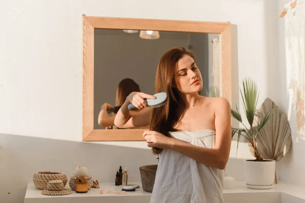 Mujer joven, sonriente y hermosa peinándose el pelo con un peine delante del espejo y sonriendo. Cuidado del cabello. — Foto de Stock