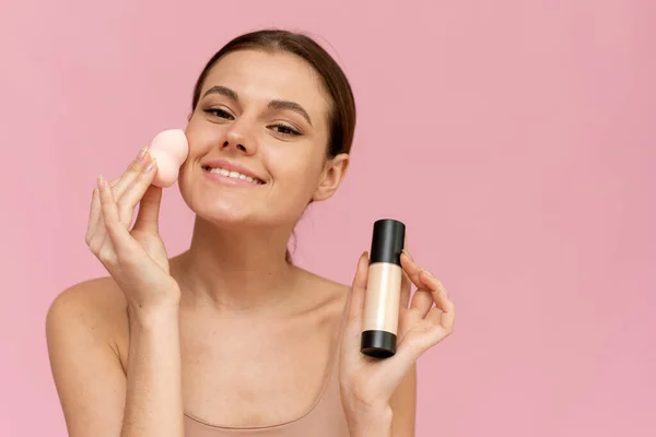 Joven hermosa mujer aplicando la fundación con esponja y mirando a la cámara en el fondo rosa. Piel perfecta, concepto de maquillaje. — Foto de Stock