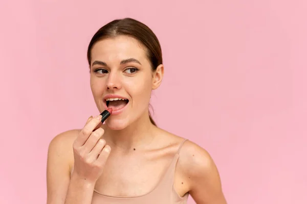 एक युवा महिला गुलाबी पृष्ठभूमि पर अपने होंठों पर नग्न लिपस्टिक लागू करती है। प्रसाधन सामग्री अवधारणा — स्टॉक फ़ोटो, इमेज