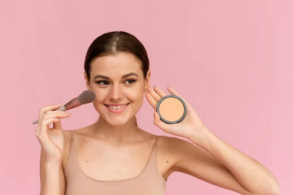 Mujer risueño aplicando polvo en su cara sobre un fondo rosa en el estudio. — Foto de Stock