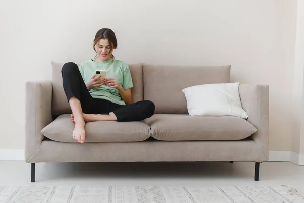 Imagen de una joven feliz y optimista sentada en casa y comprando o en las redes sociales usando el teléfono móvil en el sofá en casa. Imagen de stock