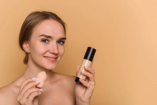 Una hermosa joven se aplica maquillaje en un fondo beige. Sonriente dama sosteniendo esponja y fundación. — Foto de Stock