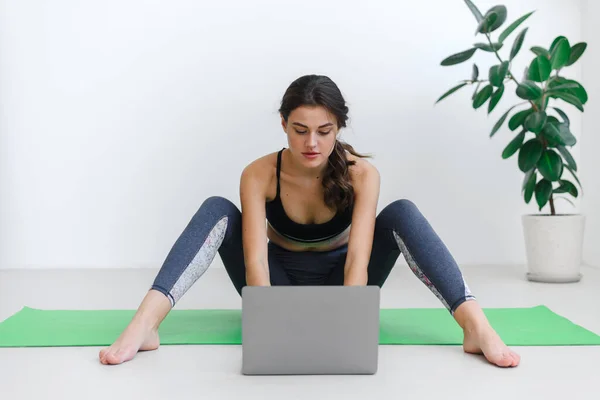 Вписується жінка робить йогу і дивиться онлайн уроки на ноутбуці, займається спортом у вітальні — стокове фото