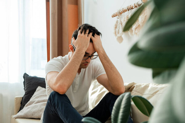 Feeling of stress, sadness of a man sitting on the couch. Frustrated, handsome, young man touching head and keeping eyes closed while sitting on sofa at home