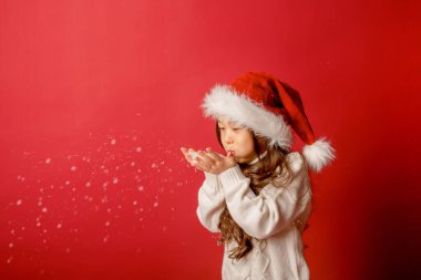 Noel Baba şapkalı Asyalı kız kırmızı arka planda kar üflüyor.