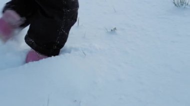 Kış, tatil, oyun, aile konsepti - yavaş çekimde bebek derin karda yürür. Çocuklar karlı yolda yürür. Soğuk mevsimde kar yağışı altında siyah tulum giymiş anaokulu çocuğu.