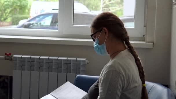 Молодая беременная женщина в медицинской маске читает на кушетке в ожидании назначения медицинского контроля в больнице. Посетите врача-гинеколога в клинике. Проверка на ожидание выздоровления живота — стоковое видео