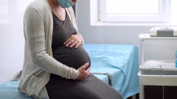 Молодая беременная женщина в медицинской защитной маске на больничном диване перед медицинской кардиотокографией. Посетите врача-гинеколога в клинике. Проверка на беременность беременной матери живота — стоковое видео