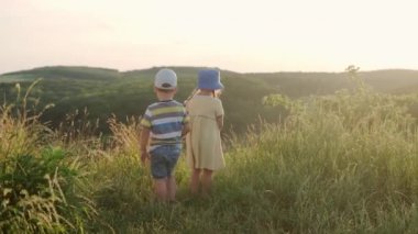 Sevimli anaokulu kızı ve oğlu gün batımından önce uzun otların arasında dağın tepesinde oturuyorlar. Vahşi çayırlarda yürüyen bir çocuk. Dağlarda mutlu bir çocuk. Çocukluk, doğa, yaşam tarzı, yaz konsepti