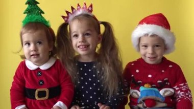 Duygusal Sevimli Mutlu Neşeli 3 Kardeş Arkadaş Bebek Kız ve Noel Baba Takım Elbiseli Çocuk sarı arka planda kameraya bakıyor. Doğum gününü kutlayan çocuk Noel Sahnesi oynuyor. Çocuk Yılbaşını Eğlenerek Geçiriyor