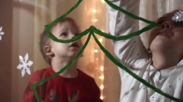 本物のかわいいお母さんと2人の小さな就学前の未成年の兄弟の女の子と男の子2-4歳は窓にクリスマスツリーを描きます。子供を持つ若い女性は雪の結晶を接着する。クリスマス、新年、冬のコンセプト. — ストック動画
