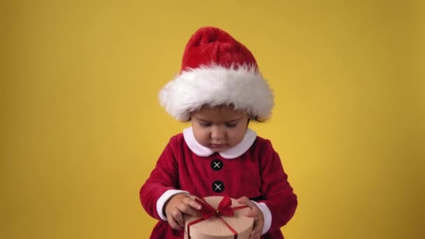 Šťastný veselý buclatý batole dítě dívka v santa oblek při pohledu na kameru na žlutém pozadí. Dítě Zahrajte si vánoční scénu Slavit narozeniny. Dítě otevřené obdržet překvapení dárkové krabice dárek Nový rok — Stock video